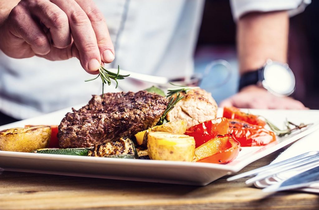 Chef adding a garnish to a plate of steak, tomatos, potatoes, and asparagus.